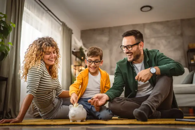 a woman, young boy, and a man holding a coin sitting on the carpet in a living room with a piggy bank in front of them