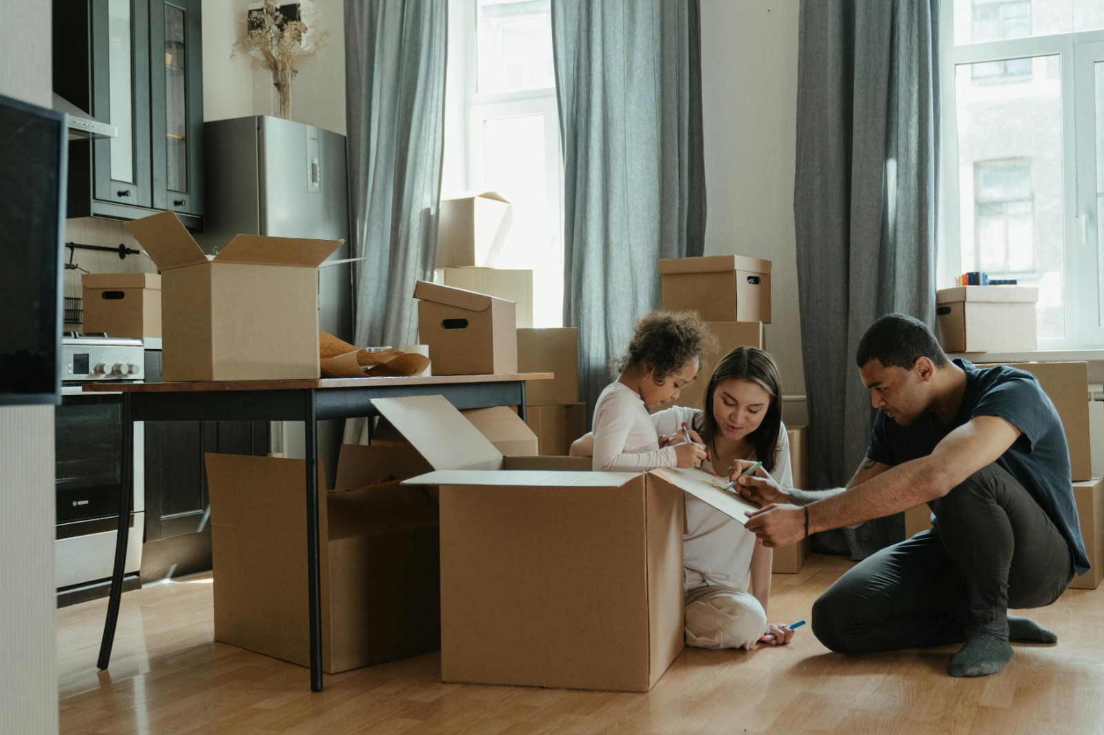 child inside a moving box with parents kneeling down next to her drawing on the box as they unpack in their new home covered by Vireya homeowners insurance