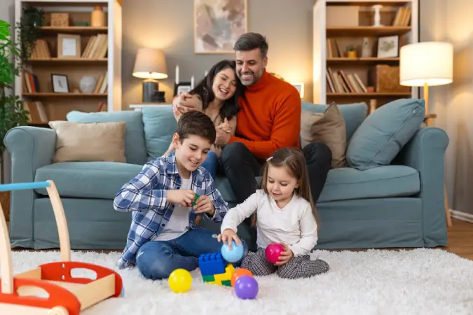 parents smiling and sitting on couch while their kids play on the rug in front of them in a home covered by homeowners insurance through Vireya