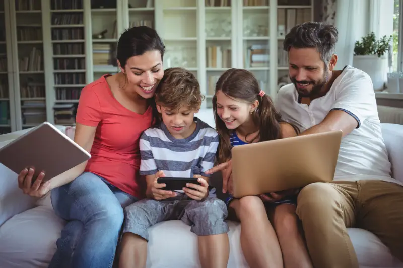 Family of four using a tablet, smartphone, and laptop together on a sofa