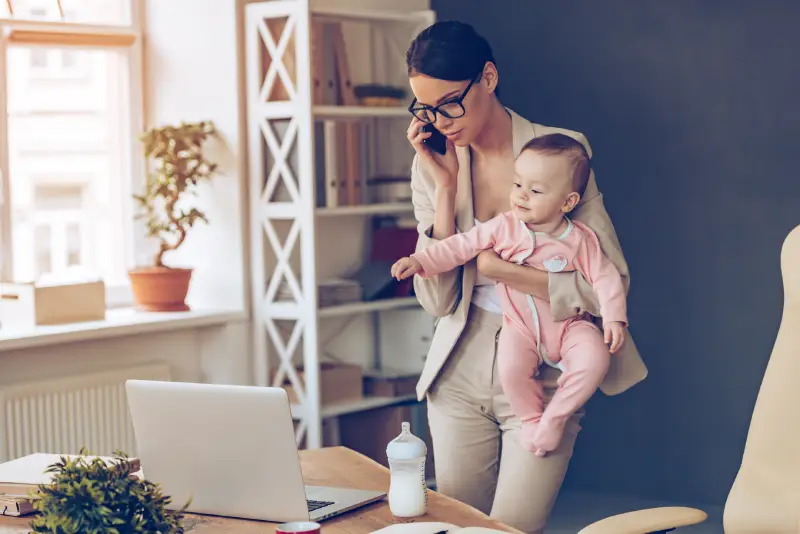 woman talking on phone with an insurance agent looking at her laptop while holding baby in her arm