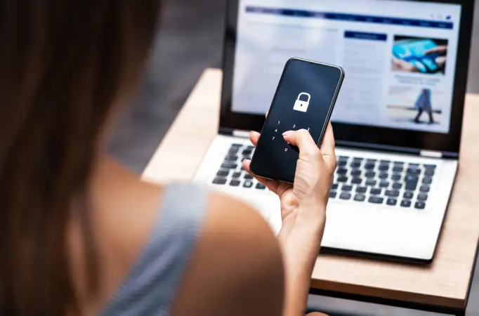 Person holding a smartphone with a lock screen, while a laptop is open in the background, symbolizing online security