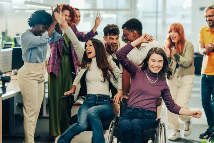 woman in an office chair next to a woman in a wheel chair with their colleagues standing behind them smiling and clapping