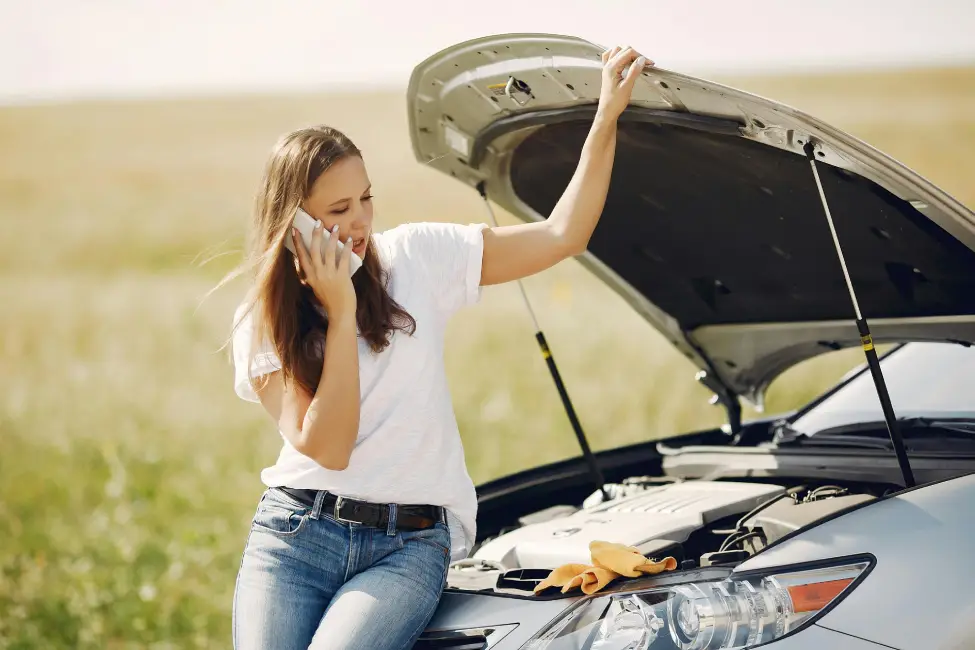 woman on the phone with roadside assistance leaning agains the front of her car with the hood open