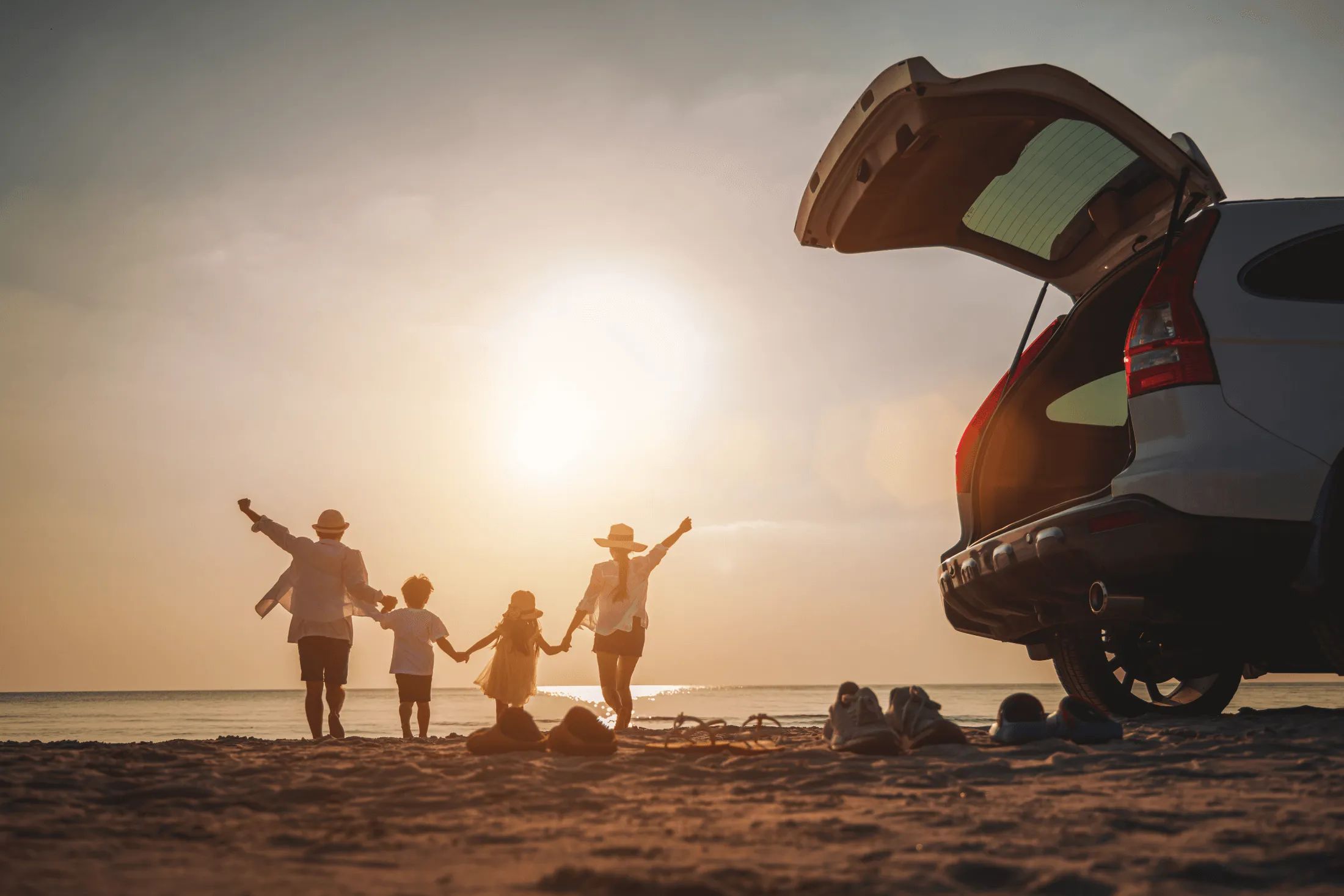 backs of a family of four all holding hands on a sunsetted beach with a car covered by Vireya auto insurance in the foreground